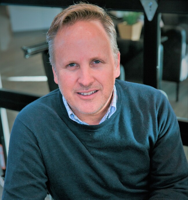 Paul Scherrer smiling and wearing a blue sweater over a checkered shirt, sitting indoors.